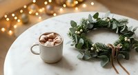 Cozy Christmas morning scene with hot chocolate topped with marshmallows and festive eucalyptus wreath on white table with warm bokeh lights.
