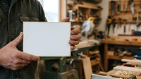 Craftsman holding blank white card in woodworking workshop with tools and wooden materials, suitable for business branding, trade services, and handmade product promotion.