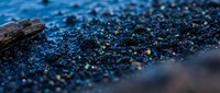 Dark volcanic black sand beach with iridescent pebbles and driftwood, shallow depth of field, moody blue tones, nature coastal texture.