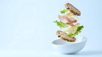 Deconstructed sandwich with flying ingredients above white bowl on clean background for food advertising and culinary design projects.
