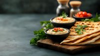 Creamy hummus served in dark bowls with pita bread triangles on wooden board, garnished with fresh parsley and cherry tomatoes against dark background.
