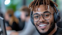 Young African American man with dreadlocks, glasses and headphones smiling in office environment, representing modern workplace diversity.