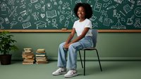 Young African American girl student sitting on chair in classroom with educational doodles on green chalkboard background for back to school concepts.