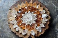 Crystal mandala arrangement with clear quartz and citrine stones in circular pattern on rustic wooden slice, creating sacred geometry design against textured gray surface.