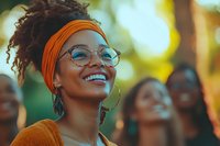 Young African American woman with natural curly hair wearing orange headscarf and glasses smiling outdoors in warm sunlight with blurred people in background.