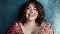 Young Caucasian woman with wavy brown hair and bright smile wearing floral dress against blue textured background, expressing joy and confidence in studio portrait.