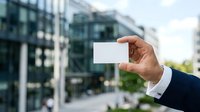 Businessman in dark suit holding blank white business card outdoors with modern office buildings in background, suitable for corporate branding and professional networking materials.