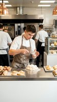 Young male baker in apron dusting pastries with powdered sugar in professional kitchen. Caucasian chef preparing baked goods at counter with team working in background.