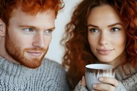 Young redhead couple sharing intimate moment with hot beverage in cozy sweaters during winter season at home together.