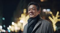 Mature Asian businessman smiling confidently at night with warm city lights creating bokeh background. Professional executive portrait outdoors.