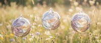 Three delicate glass ornaments hanging in golden sunlit meadow with wildflowers creating dreamy bokeh background for seasonal decoration concepts.