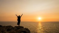 Woman celebrating freedom with raised arms on rocky cliff overlooking ocean at golden sunset, expressing joy and achievement in nature.