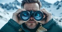 Bearded Caucasian man in winter jacket using binoculars against snowy mountain backdrop, with mountain reflection visible in binocular lenses creating dramatic outdoor scene.