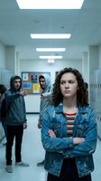 Teenage girl with curly hair stands confidently in school hallway while male students watch from background, depicting bullying or social dynamics.