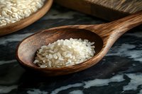 Raw white rice grains in rustic wooden spoon on dark marble background, close-up view showing texture and natural grain details in warm lighting.