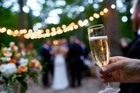 Champagne glass held at outdoor wedding reception with string lights and blurred couple in background. Celebration toast at evening garden party.