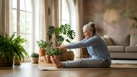 Senior woman practicing yoga stretching exercise on mat in bright living room with houseplants creating peaceful wellness atmosphere at home.