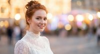 Young redhead woman in white lace dress smiling at camera with warm bokeh city lights background during golden hour for lifestyle and fashion content.