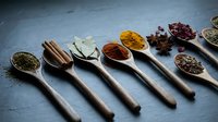 Aromatic spices and herbs arranged in wooden spoons on dark slate background for culinary cooking and seasoning recipes.
