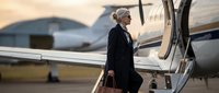 Senior woman in business attire boarding private jet at airport runway. Elegant traveler with luggage preparing for executive flight departure.