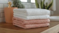 Stack of soft cotton towels in white and coral pink colors on wooden surface in modern bathroom with plants and soap dispenser creating spa atmosphere.