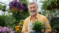 Cheerful senior man in yellow polo shirt and beige apron holding potted fern plant at garden center, surrounded by colorful flowers and hanging baskets.