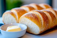 Freshly baked braided bread loaf with golden brown crust served with butter pat in white ceramic bowl, close-up view showing soft texture and glossy surface.