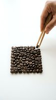 Hand using wooden tweezers to arrange roasted coffee beans in a square shape on a white background, minimalist food concept.