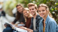 Diverse group of young college students studying outdoors on campus, smiling at camera while surrounded by greenery.