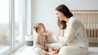 Young Caucasian mother in cream sweater bonding with toddler daughter in nursery room with wooden crib and natural light from window for family lifestyle.