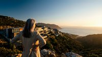 Woman with long dark hair wearing casual clothing stands on hillside overlooking coastal city at sunset, gazing at urban landscape and ocean view during golden hour.
