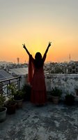 Woman in red dress celebrating victory with raised arms on urban rooftop terrace at golden sunset over city skyline with communication towers and buildings.