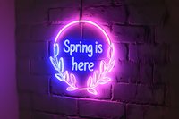 Glowing neon sign reading Spring is here with floral wreath elements mounted on brick wall, illuminated in pink and blue light creating modern seasonal ambiance.