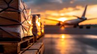 Cargo pallets stacked at airport during dramatic orange sunset with commercial airplane silhouette in background, reflecting on wet tarmac surface.