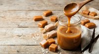 Homemade salted caramel sauce dripping from wooden spoon into glass jar with vanilla beans and caramel candies on rustic wooden background for desserts.