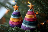 Handmade knitted Christmas trees with colorful striped patterns and star toppers displayed among evergreen branches with warm holiday lights creating festive atmosphere.