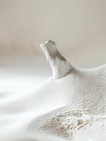 Minimalist still life composition featuring bleached driftwood piece emerging from soft white sand dunes, creating gentle shadows and textures in natural light.