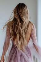 Young woman with flowing blonde balayage hair wearing romantic sheer lavender dress with open back, captured in soft natural light against white wall.