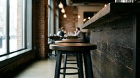Modern industrial cafe interior with wooden bar counter and black metal stool near brick wall and large window, featuring blurred background with customers and warm lighting for restaurant or hospitality design.