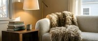 Cozy living room corner with beige armchair, faux fur throw blanket, wooden side table, lamp, books, and cup near bright window for home decor.