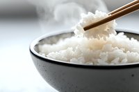 Cooked white rice in gray ceramic bowl with wooden chopsticks picking up a portion, ready for Asian cuisine preparation and dining.