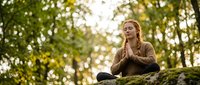 Young blonde woman practicing meditation outdoors in peaceful forest setting with hands in prayer position on moss-covered log during golden hour sunlight.