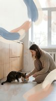 Young woman in beige sweater feeding tabby cat in modern kitchen with abstract wall art and natural light streaming through window.