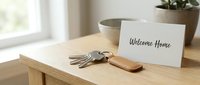 House keys with leather keychain and Welcome Home card on wooden table, bright interior background, real estate and moving concept.