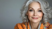 Mature woman with silver gray wavy hair and natural makeup wearing bright orange clothing smiles warmly against neutral gray background in horizontal banner format.