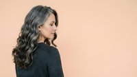 Middle-aged woman with long wavy dark hair and gray streaks, wearing dark sweater, shown in profile against neutral beige background. Suitable for lifestyle, beauty, and wellness content.