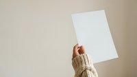 Cropped hand in beige knit sweater holding blank white paper sheet against neutral background, suitable for mockups, presentations, advertising campaigns, and promotional materials.
