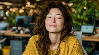 Young woman with curly brown hair and closed eyes wearing yellow sweater in peaceful meditation pose at modern office workspace with plants.