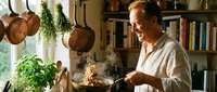 Middle-aged Caucasian man cooking in rustic kitchen with copper pots, herbs, and books. Chef preparing meal at home with traditional cookware and natural ingredients.