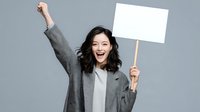 Excited young Asian woman holding blank white protest sign with raised fist celebrating victory or activism on gray background for social campaigns.
